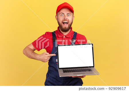 Portrait of excited bearded handyman in overall holding laptop with empty display, pointing at screen and looking at camera with open mouth. Indoor studio shot isolated on yellow background. 92909617