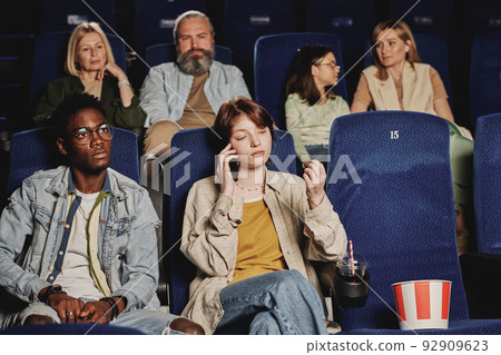 Young gen Z Caucasian woman talking on phone while watching movie at cinema distracting other people Young gen Z Caucasian woman talking on phone while watching movie at cinema distracting other people 92909623