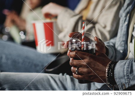 Selective focus shot of unrecognizable Black man holding plastic cup with cola drink at cinema Selective focus shot of unrecognizable Black man holding plastic cup with cola drink at cinema 92909675