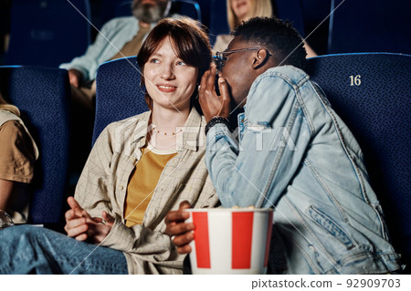 Young Black man whispering something in his Caucasian girlfriend ear while watching movie at cinema 92909703