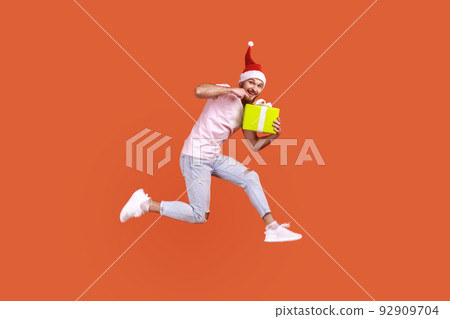 Portrait of attractive man in santa hat jumping in air and pointing at present box in his hands, looking at camera, wearing pink T-shirt and jeans. Indoor studio shot isolated on orange background. 92909704