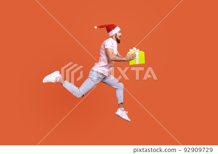 Bearded man jumping in air or running quickly fast to give present box for Christmas, expressing happiness, wearing pink T-shirt and santa hat. Indoor studio shot isolated on orange background. Bearded man jumping in air or running quickly fast to give present box for Christmas, expressing happiness, wearing pink T-shirt and santa hat. Indoor studio shot isolated on orange background. 92909729