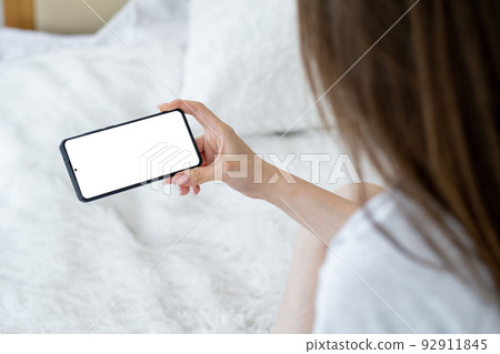 Virtual life. Digital mockup. Online connection. Unrecognizable relaxed woman holding smartphone with blank screen laying bed in light room interior. 92911845