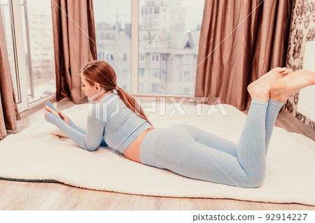 Side view portrait of relaxed woman with long hair lying on carpet at home. She is dressed in a blue tracksuit, holding a phone in her hands. Side view portrait of relaxed woman with long hair lying on carpet at home. She is dressed in a blue tracksuit, holding a phone in her hands. 92914227