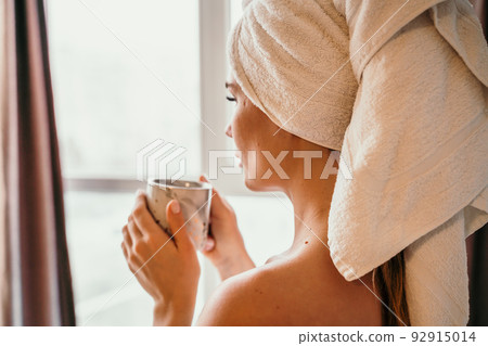 Young serene relaxed woman in spa bath towel drinking hot beverage tea coffee after taking shower bath at home. Beauty treatment, hydration concept. 92915014