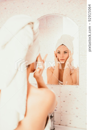 Young beautiful woman using face cream moisturizing lotion after bath. Pretty attractive girl in a towel on her head stands in front of a mirror in a home bathroom. Daily hygiene and skin care 92915064