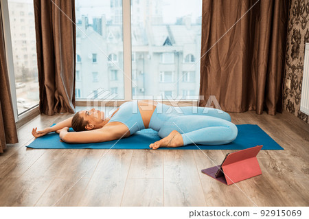 Young athletic attractive woman practicing yoga doing exercises. Works out at home or in a yoga studio, sportswear, blue pants and a full-length top indoors. Healthy lifestyle concept Young athletic attractive woman practicing yoga doing exercises. Works out at home or in a yoga studio, sportswear, blue pants and a full-length top indoors. Healthy lifestyle concept 92915069