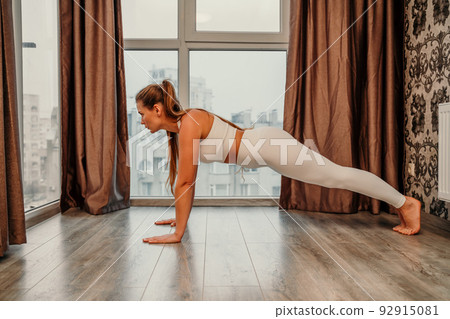 Young attractive athletic woman in white sportswear doing push ups or pushups on gym mat at home, practicing yoga plank pose, exercising indoors, healthy active lifestyle concept Young attractive athletic woman in white sportswear doing push ups or pushups on gym mat at home, practicing yoga plank pose, exercising indoors, healthy active lifestyle concept 92915081