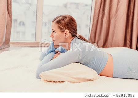 Side view portrait of relaxed woman with long hair lying on carpet at home. She is wearing a blue tracksuit. 92915092