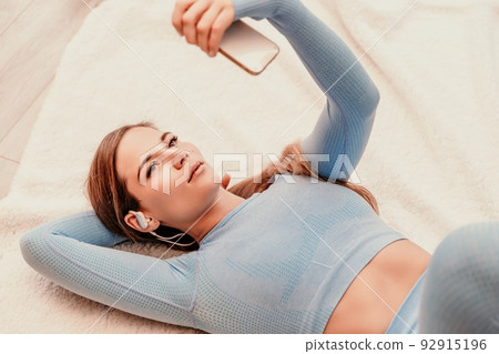 Top view portrait of relaxed woman listening to music with headphones lying on carpet at home. She is dressed in a blue tracksuit. Top view portrait of relaxed woman listening to music with headphones lying on carpet at home. She is dressed in a blue tracksuit. 92915196