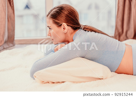 Side view portrait of relaxed woman with long hair lying on carpet at home. She is dressed in a blue tracksuit, holding a phone in her hands. Side view portrait of relaxed woman with long hair lying on carpet at home. She is dressed in a blue tracksuit, holding a phone in her hands. 92915244