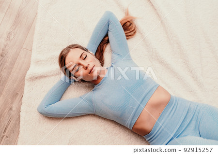 Top view portrait of relaxed woman listening to music with headphones lying on carpet at home. She is dressed in a blue tracksuit. 92915257
