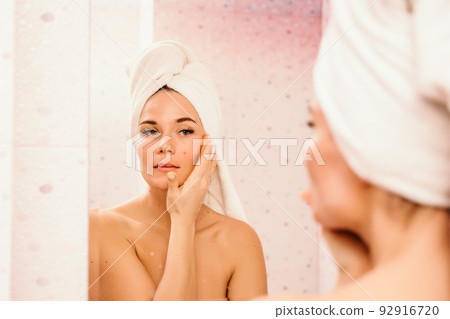 Young beautiful woman using face cream moisturizing lotion after bath. Pretty attractive girl in a towel on her head stands in front of a mirror in a home bathroom. Daily hygiene and skin care Young beautiful woman using face cream moisturizing lotion after bath. Pretty attractive girl in a towel on her head stands in front of a mirror in a home bathroom. Daily hygiene and skin care 92916720