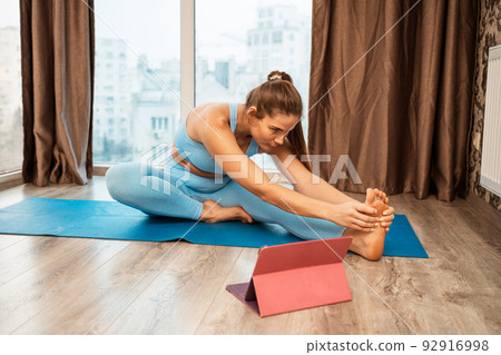 Young athletic attractive woman practicing yoga doing exercises. Works out at home or in a yoga studio, sportswear, blue pants and a full-length top indoors. Healthy lifestyle concept Young athletic attractive woman practicing yoga doing exercises. Works out at home or in a yoga studio, sportswear, blue pants and a full-length top indoors. Healthy lifestyle concept 92916998