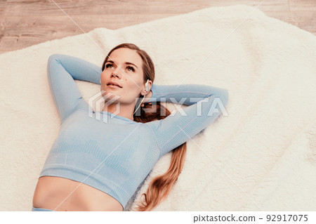 Top view portrait of relaxed woman listening to music with headphones lying on carpet at home. She is dressed in a blue tracksuit. Top view portrait of relaxed woman listening to music with headphones lying on carpet at home. She is dressed in a blue tracksuit. 92917075