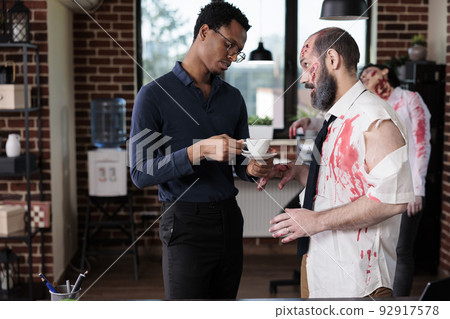African american adult chatting with zombie in business office, undead spooky corpse chatting with person in startup workplace. Evil horror macabre monster with bloody scars and decayed. 92917578