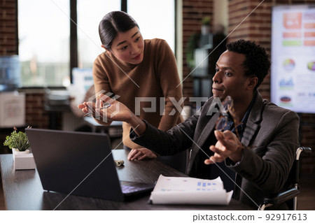 African american wheelchair user working with asian woman on business project in disability friendly office. Paralyzed man with chronic impairment doing teamwork with colleague. 92917613