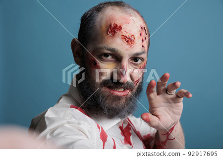 Halloween zombie taking picture on smartphone, looking crazy evil and aggressive in studio. Undead brain eating monster using camera to take photo on mobile phone, being creepy and frightening. 92917695