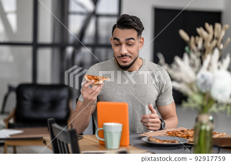 Man eating pizza looking in surprise at tablet Man eating pizza looking in surprise at tablet 92917759