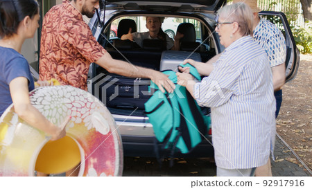 Happy people loading vehicle with luggage and trolleys while going on weekend citybreak together. Big family packing voyage luggage in car trunk while getting ready for summer field trip. 92917916