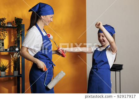 Mother and child having fun painting house walls with orange paint, using paintbrush and roller to redecorate apartment room and enjoy renovation. Woman with kid decorating home. 92917917