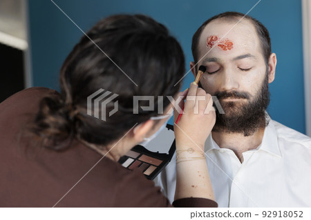 Artist using makeup effects to create zombie costume and scary dramatic corpse look. Man looking deceased and infected after having bloody scars and monster face, creepy design. Artist using makeup effects to create zombie costume and scary dramatic corpse look. Man looking deceased and infected after having bloody scars and monster face, creepy design. 92918052