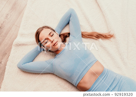 Top view portrait of relaxed woman listening to music with headphones lying on carpet at home. She is dressed in a blue tracksuit. 92919320