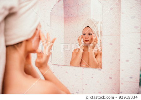 Young beautiful woman using a moisturizing facial mask after taking a bath. Pretty attractive girl in a towel on her head stands in front of a mirror in a home bathroom. Daily hygiene and skin care 92919334