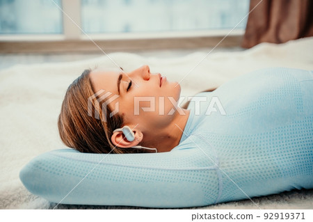 Side view portrait of relaxed woman listening to music with headphones lying on carpet at home. She is dressed in a blue tracksuit. 92919371