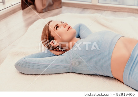 Top view portrait of relaxed woman listening to music with headphones lying on carpet at home. She is dressed in a blue tracksuit. Top view portrait of relaxed woman listening to music with headphones lying on carpet at home. She is dressed in a blue tracksuit. 92919400