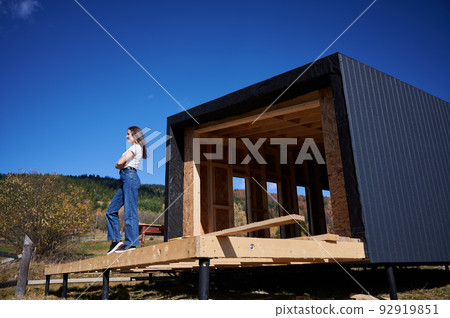 Happy woman on construction site standing on terrace at unfinished wooden frame house in the Scandinavian style barnhouse on sunny day. 92919851