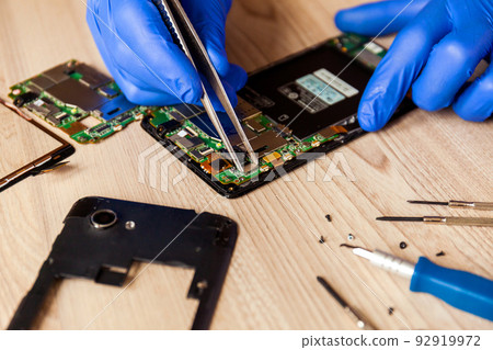 The technician repairing the smartphone's motherboard in the workshop on the table. 92919972