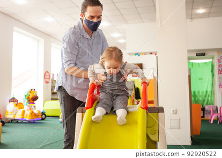 young Dad in a medical mask rolls his daughter from the slide in the game room. Child development center, kindergarten, fun pastime. Family together 92920522