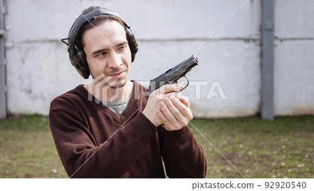 A young man is aiming for a gun. A man wearing protective headphones. outdoor tyre shooting range. hobby. firearm against the white wall A young man is aiming for a gun. A man wearing protective headphones. outdoor tyre shooting range. hobby. firearm against the white wall 92920540