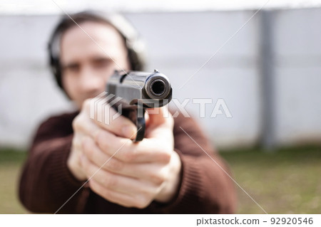 A young man is aiming for a gun. A man wearing protective headphones. outdoor tyre shooting range. hobby. firearm against the white wall copy space A young man is aiming for a gun. A man wearing protective headphones. outdoor tyre shooting range. hobby. firearm against the white wall copy space 92920546