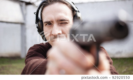 A young man is aiming for a gun. A man wearing protective headphones. outdoor tyre shooting range. hobby. firearm against the white wall close up 92920550