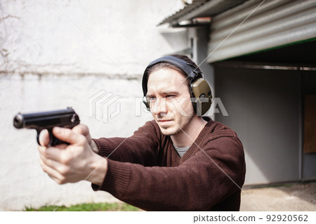 A young man is aiming for a gun. A man wearing protective headphones. outdoor tyre shooting range. hobby. firearm against the white wall A young man is aiming for a gun. A man wearing protective headphones. outdoor tyre shooting range. hobby. firearm against the white wall 92920562