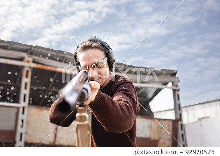A young man in protective glasses and headphones. A pump-action shotgun. Tyre's outdoors. A wall and a roof with bullet holes. Sport close up A young man in protective glasses and headphones. A pump-action shotgun. Tyre's outdoors. A wall and a roof with bullet holes. Sport close up 92920573