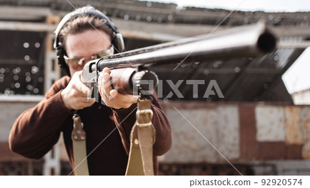 A young man in protective glasses and headphones. A pump-action shotgun. Tyre's outdoors. A wall and a roof with bullet holes. Sport A young man in protective glasses and headphones. A pump-action shotgun. Tyre's outdoors. A wall and a roof with bullet holes. Sport 92920574