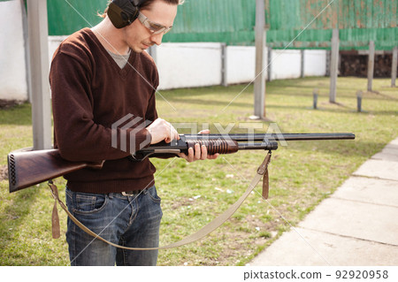 A young man charges a pump-action shotgun with a Ammo. 12 caliber. Tyre outdoor. A man in headphones and goggles is preparing to shoot. Firearms for sports shooting, hobby. A young man charges a pump-action shotgun with a Ammo. 12 caliber. Tyre outdoor. A man in headphones and goggles is preparing to shoot. Firearms for sports shooting, hobby. 92920958