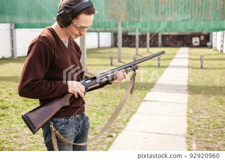 A young man charges a pump-action shotgun with a Ammo. 12 caliber. Tyre outdoor. A man in headphones and goggles is preparing to shoot. Firearms for sports shooting, hobby. 92920960