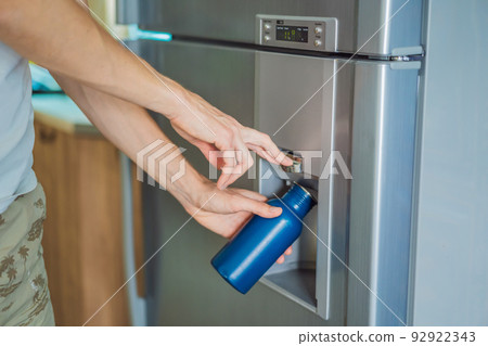 Male hand is pouring cold water and ice cubes in a metal bottle from dispenser of home fridge 92922343