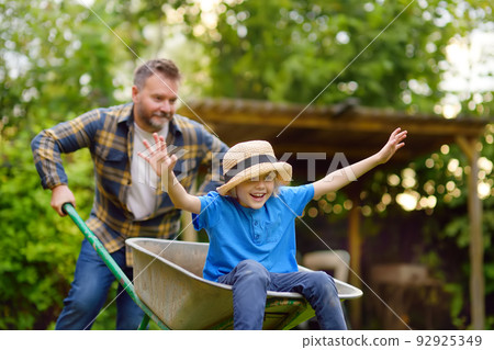 Happy little boy having fun in a wheelbarrow pushing by dad in domestic garden on warm sunny day. Active outdoors games for family with kids in the backyard in summer Happy little boy having fun in a wheelbarrow pushing by dad in domestic garden on warm sunny day. Active outdoors games for family with kids in the backyard in summer 92925349