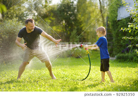 Funny little boy with his father playing with garden hose in sunny backyard. Preschooler child having fun with spray of water. Summer outdoors activity for family with kids. Funny little boy with his father playing with garden hose in sunny backyard. Preschooler child having fun with spray of water. Summer outdoors activity for family with kids. 92925350