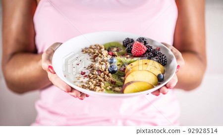 Yogurt, blackberry muesli, raspberries, blueberries, kiwi and peaches in a white bowl holding young woman 92928514