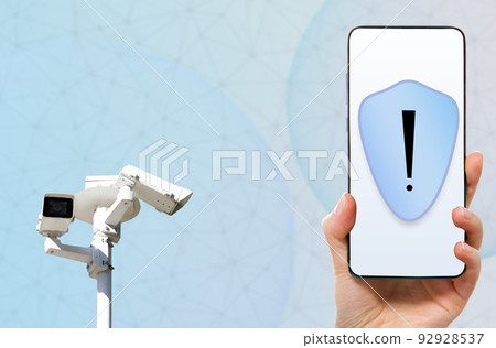 Supervision app and monitoring. Female hand holds a smartphone showing shield at screen. Surveillance camera at the background with copy space. Concept of CCTV and integrated video control system Supervision app and monitoring. Female hand holds a smartphone showing shield at screen. Surveillance camera at the background with copy space. Concept of CCTV and integrated video control system 92928537