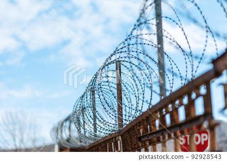 A metal fence with barbed wire under tension. Bright blue sky on the background. The concept of border protection, prison and military base A metal fence with barbed wire under tension. Bright blue sky on the background. The concept of border protection, prison and military base 92928543