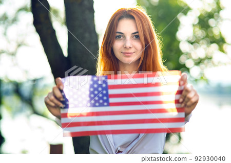 Happy young red haired woman holding USA national flag in her hands outdoors in summer. Positive girl celebrating United States independence day. 92930040