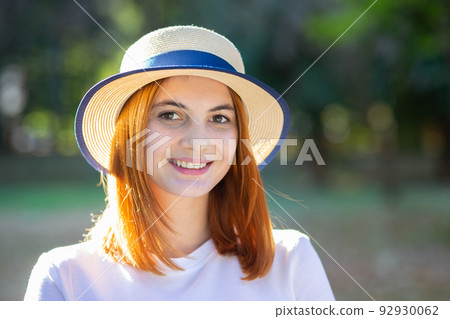 Closeup portrait of redhead hipster teenage girl in yellow hat smiling outdoors in sunny summer park. 92930062
