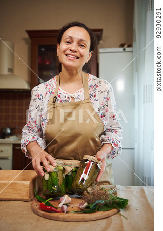 Charming multi-ethnic woman, housewife stands in the home kitchen interior and smiles looking at camera, showing jars with homemade pickled cucumbers and chili peppers. Seasonal canning of vegetables Charming multi-ethnic woman, housewife stands in the home kitchen interior and smiles looking at camera, showing jars with homemade pickled cucumbers and chili peppers. Seasonal canning of vegetables 92930291